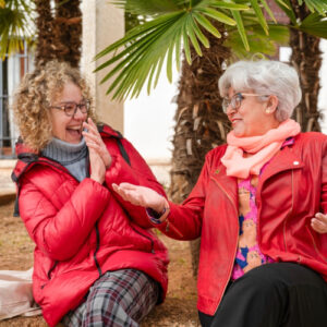 Two older women in red jackets sit outdoors under a palm tree, laughing and chatting together. One has curly hair and glasses, the other has short gray hair and a pink scarf.