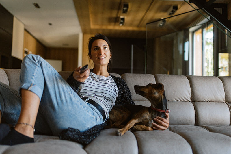 Woman relaxing on a beige sofa with a small dog. Shes wearing jeans and a striped shirt, holding a TV remote, and looking forward. The room has modern decor with wooden ceilings and a large window letting in natural light.