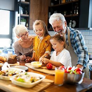 Two smiling grandparents and two young girls prepare food together in a bright kitchen, surrounded by dishes, vegetables, and orange juice on the counter. The family appears happy and engaged in cooking.