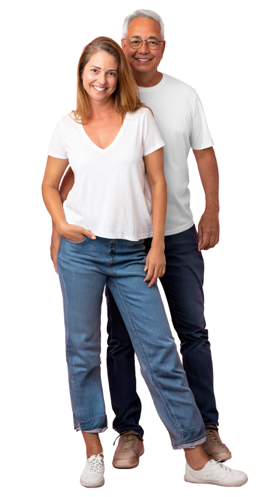 A smiling woman and an older man, who may experience hearing loss, stand together against a white background. Both wear casual outfits: white tops and blue jeans. The woman stands slightly in front, with her hand in her pocket.